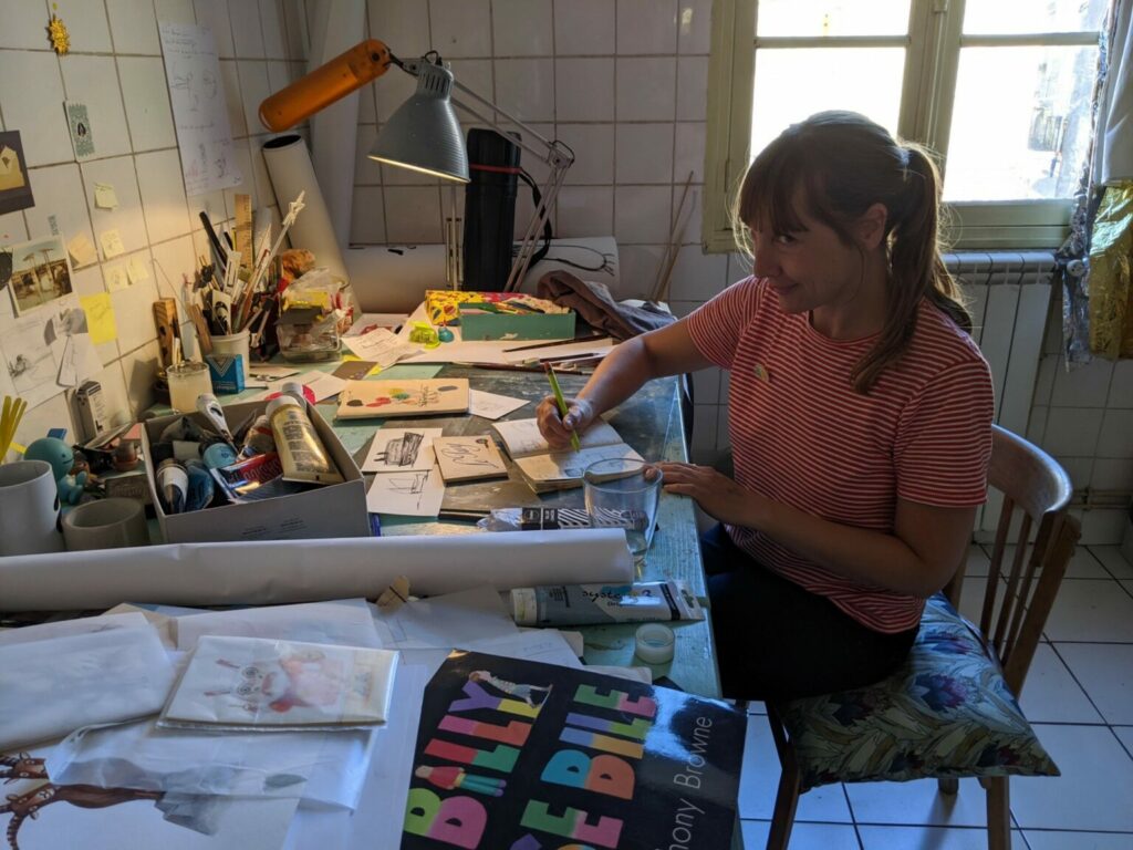 A woman with a ponytail in a red and white striped shirt sits at a messy desk. Light shines in from the window behind her. She looks up at the camera, with her hands on the desk holding a pen and small notebook. The desk is littered with books, papers, and art supplies. 