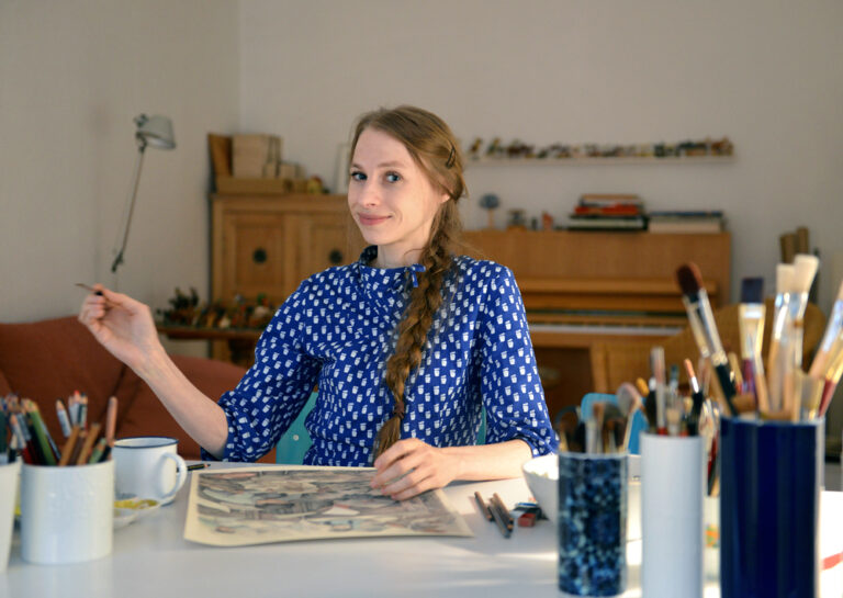 A young woman in a blue blouse holds a paintbrush and smiles for the photo. She has a painting in progress on the desk in front of her, and cups and holders frame her, filled with paint brushes and drawing tools. 